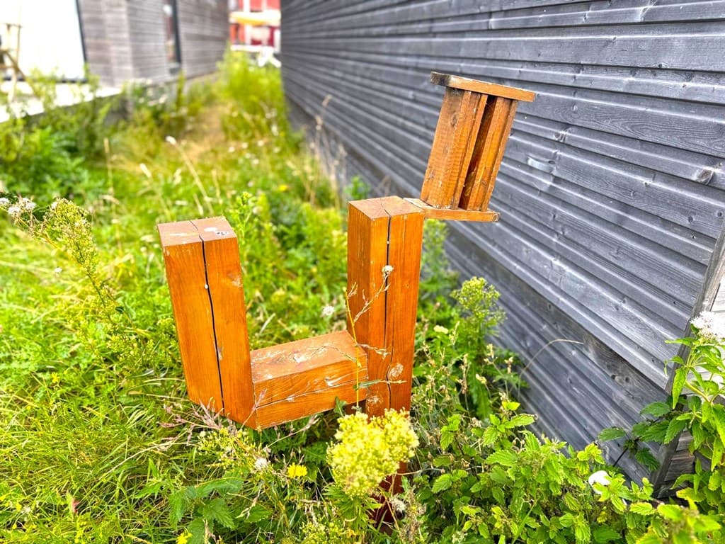 Houten constructie in een groene, ongerepte tuin met gras en planten rondom, naast een houten wand.