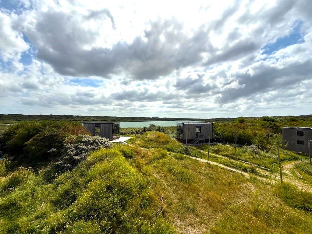 Uitzicht op de waterkant met groene begroeiing en enkele eenvoudige woningen in de verte onder een bewolkte lucht