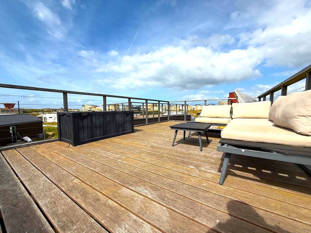 Ruime houten dakterras met loungezitplaatsen en uitzicht op de omgeving en blauwe lucht met enkele wolken