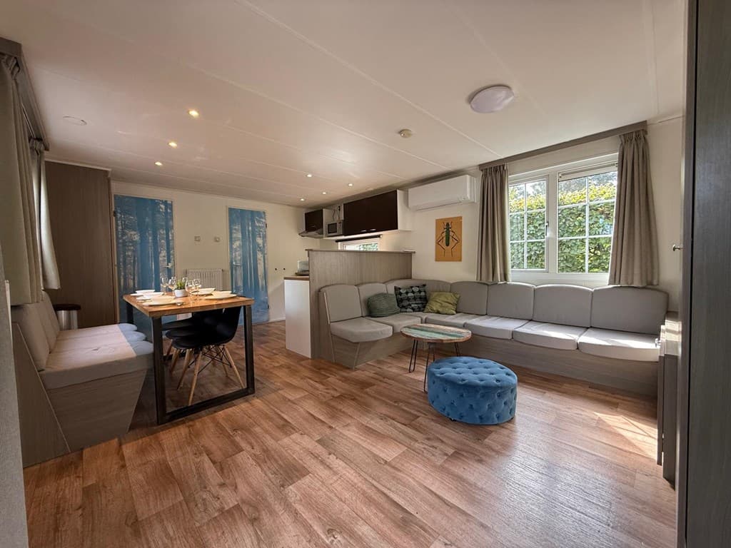 Interieur van een woning met een zithoek, eettafel en keuken, grote ramen met uitzicht naar buiten en houten vloeren.