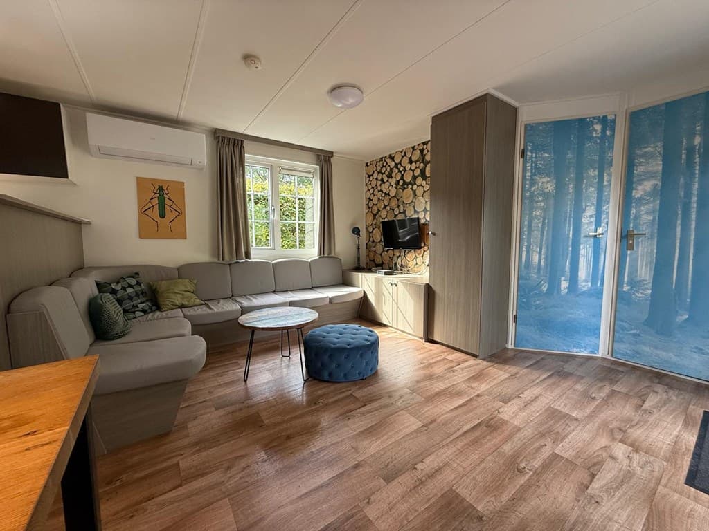 Woonkamer met een L-vormige bank, een ronde tafel, TV en glazen deuren met een bosafdruk.