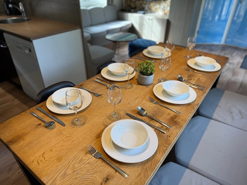 Eettafel met zes borden, glazen en bestek, omringd door zithoek en moderne inrichting in lichte kleuren.