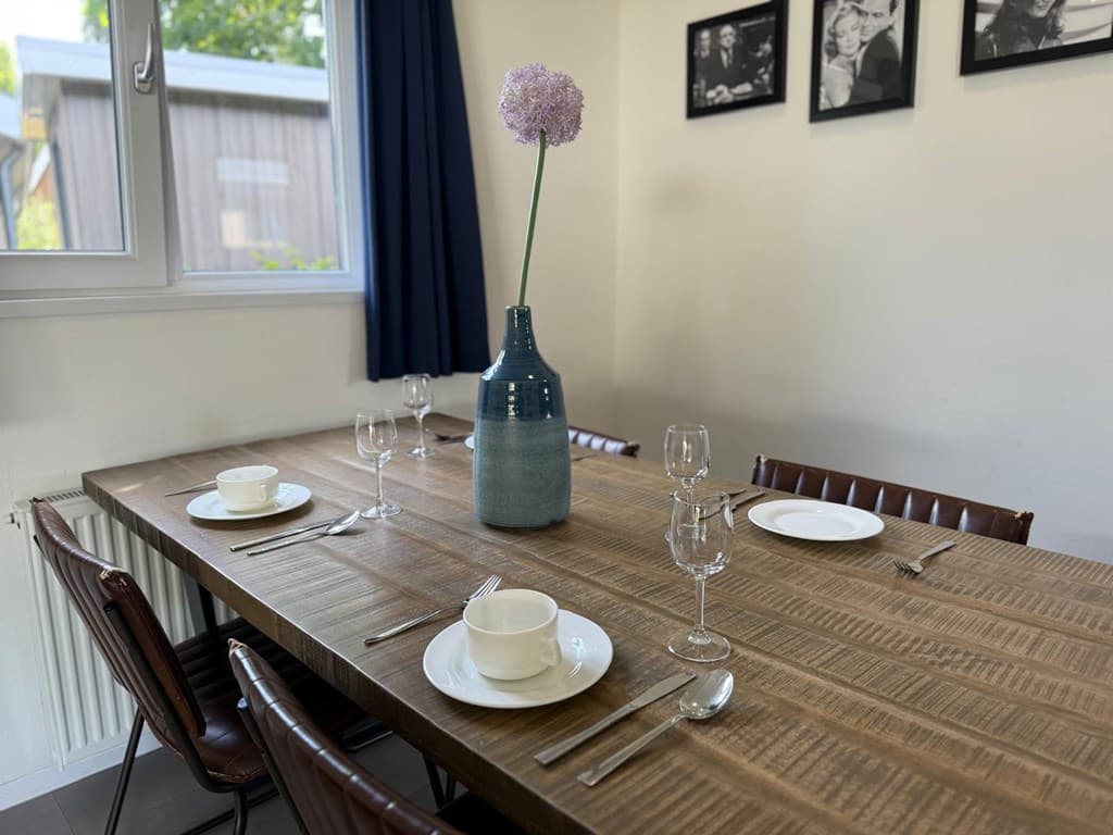 Eetkamertafel met vier stoelen, gedekt met servies, glazen en een vaas met bloem, tegen een lichte muur