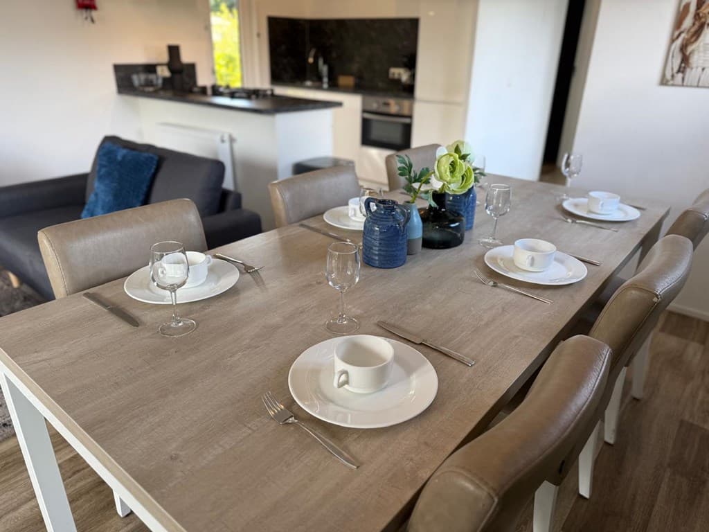 Eetkamer met een lange tafel, zes stoelen, gedekte borden en zicht op de open keuken met moderne apparatuur.