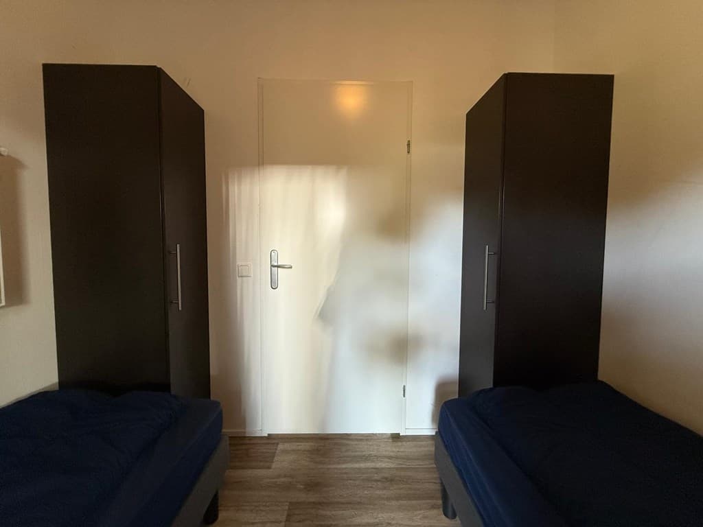 Kamer met twee eenpersoonsbedden en kastjes aan weerszijden, deur achterin zichtbaar.