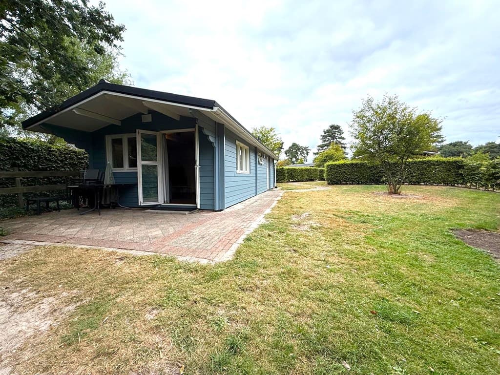 Blauw houten huisje met terras en tuin in Resort Arcen, omgeven door groene hagen en bomen.