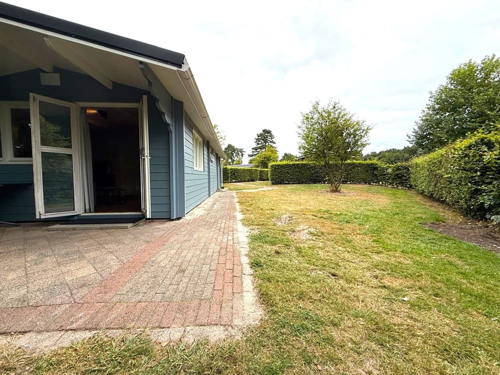 Huis met een betegeld terras en een grasveld omringd door groene hagen en struiken.