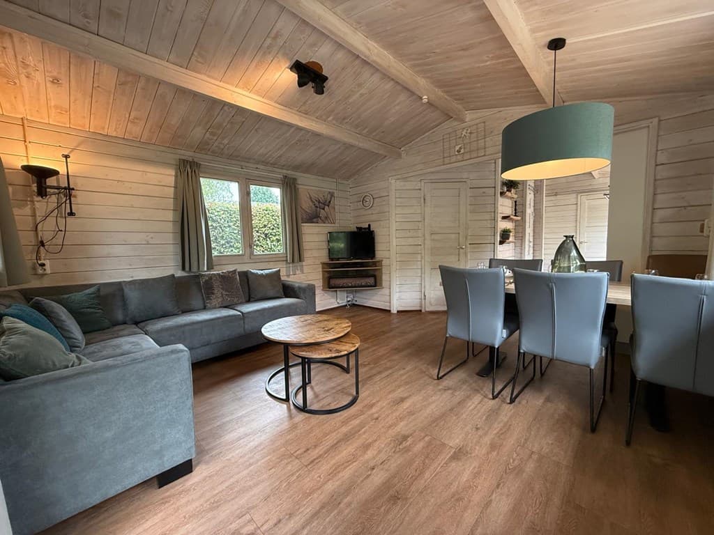 Houten woonkamer met bank, eettafel en televisie, lichte muren en plafond met houten balken.