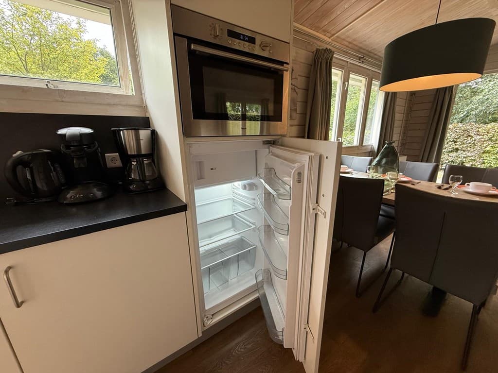 Open koelkast in een moderne keuken met koffiezetapparaat en eettafel met stoelen op de achtergrond.
