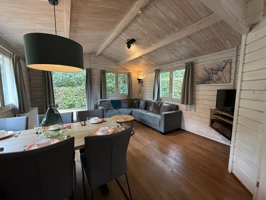 Woonkamer met houten balken, grote eettafel, zithoek en televisie, omgeven door ramen met uitzicht op groen.