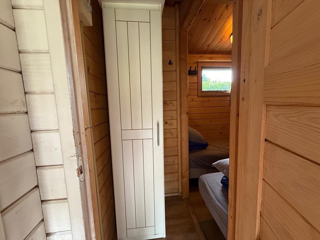 In een houten gang staat een witte kast naast een deur naar een slaapkamer met twee bedden.