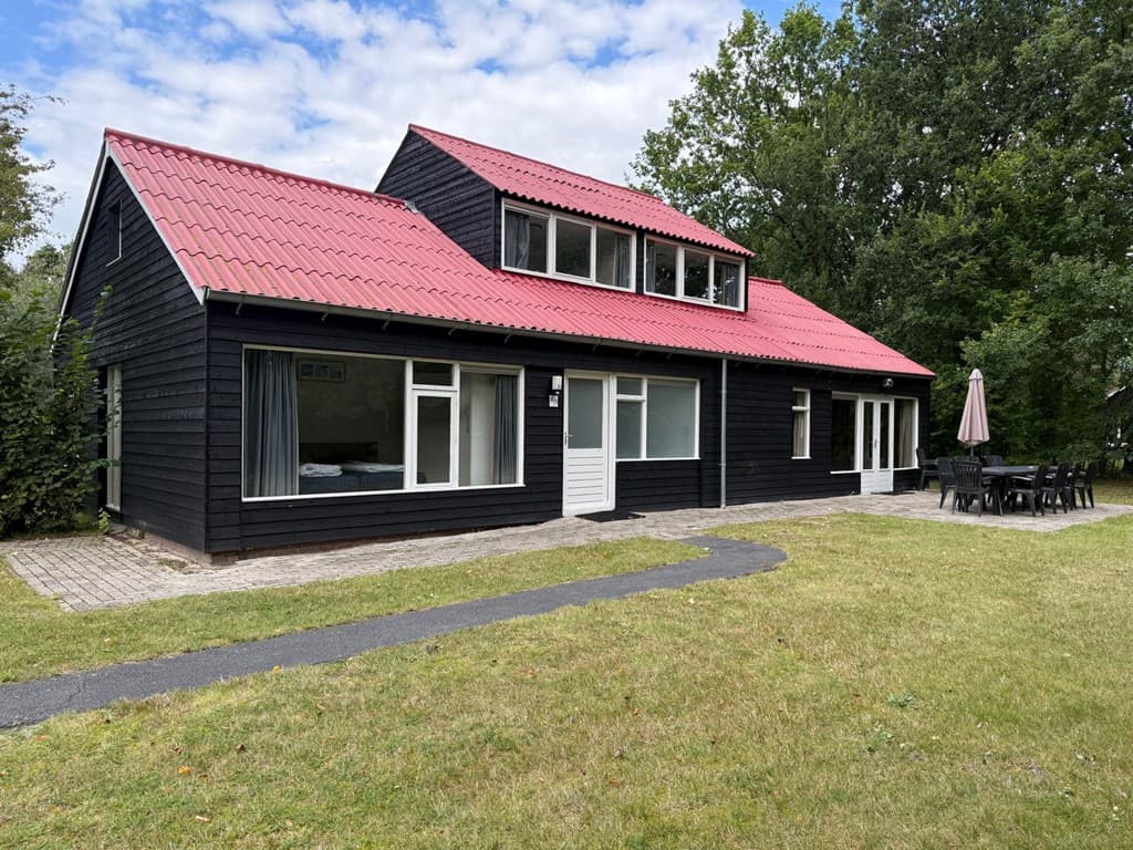 Zwart houten vakantiehuis met rode dakbedekking, grote ramen en terras met tuintafel, omgeven door groen.
