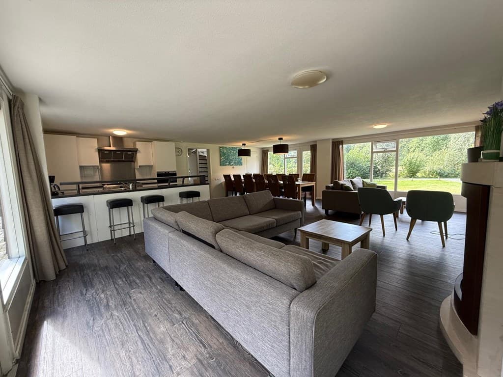 Zithoek met een grote bank en stoelen, open keuken met bar en grote eettafel, uitzicht op de tuin door brede ramen