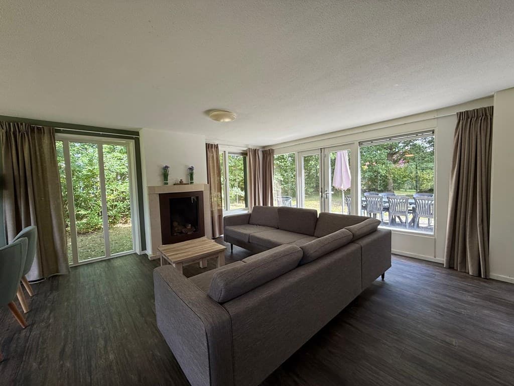 Woonkamer met een bank, haard en grote ramen die uitzicht bieden op een terras met tuinmeubilair.