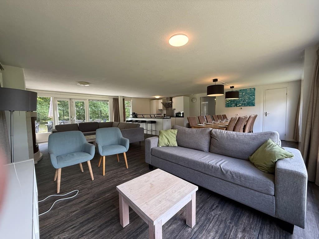 Ruime woonkamer met een grijze bank, twee blauwe stoelen en een houten salontafel, zicht op een open keuken en eettafel.