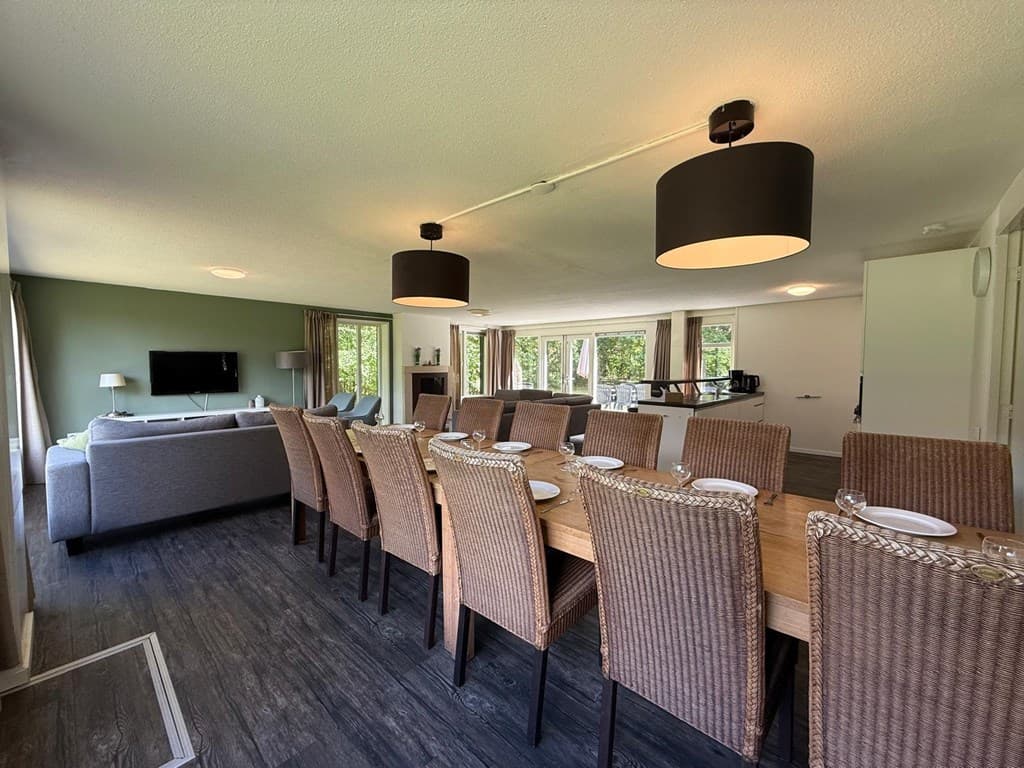 Ruime eetkamer met lange tafels, comfortabele stoelen en een zithoek met bank en tv, met uitzicht op groene natuur.