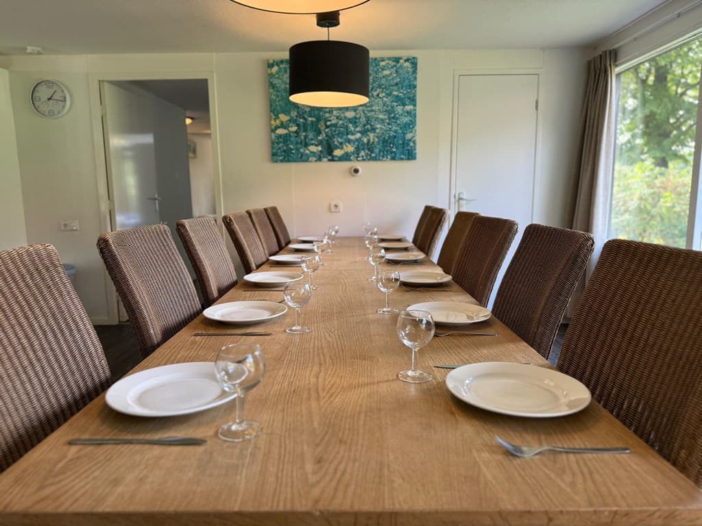 Eettafel met twaalf stoelen, gedekte borden en glazen in een lichte ruimte met uitzicht naar buiten.