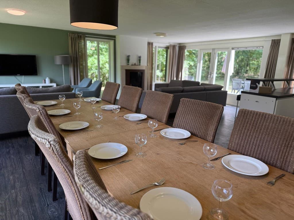 Eetkamer met lange houten tafel, stoelen en glazen, zicht op comfortabele zithoek en grote ramen naar buiten.