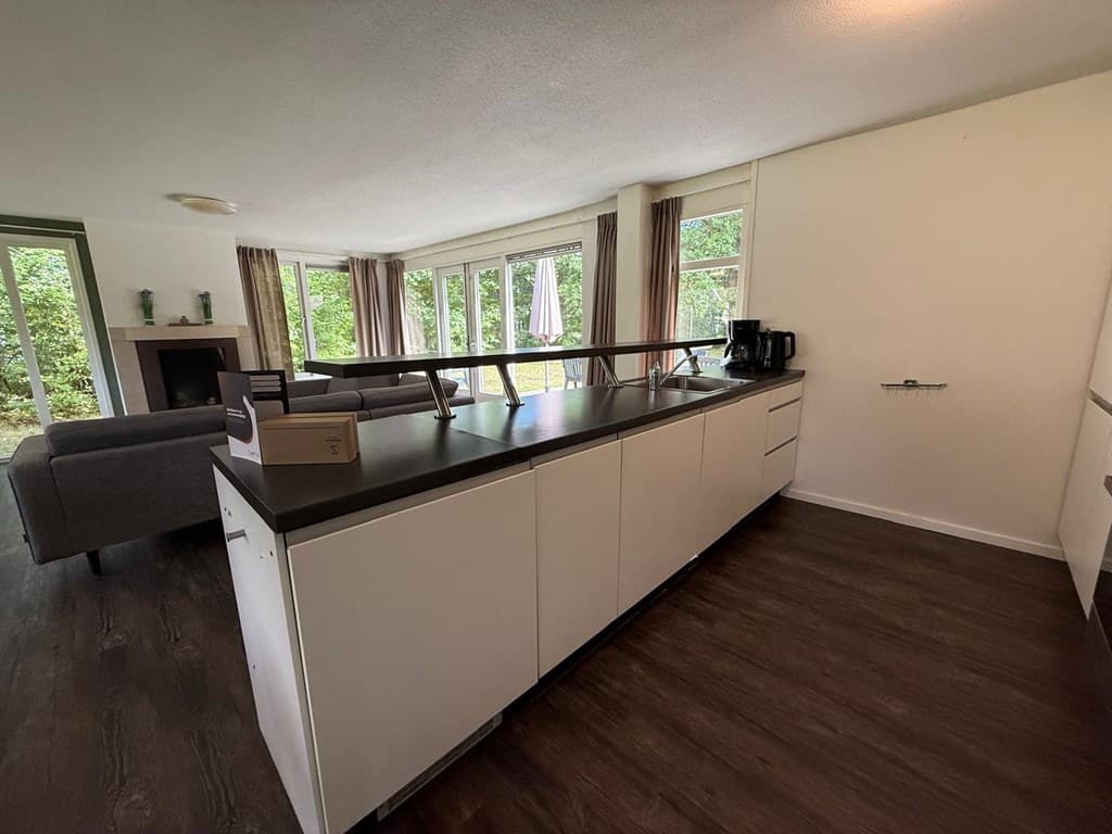 Keuken met een kookeiland en uitzicht op groene tuin door grote ramen, zithoek zichtbaar achterin de ruimte