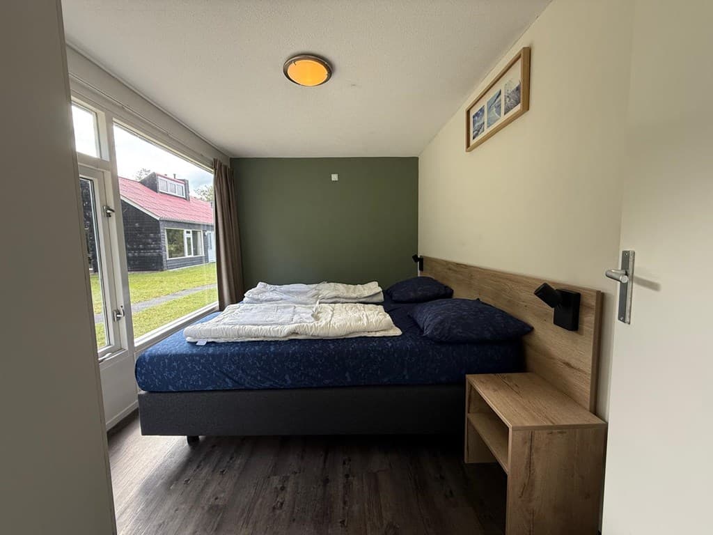 Slaapkamer met dubbel bed, nachtkastje en raam met uitzicht op een groene buitenruimte en andere woningen.