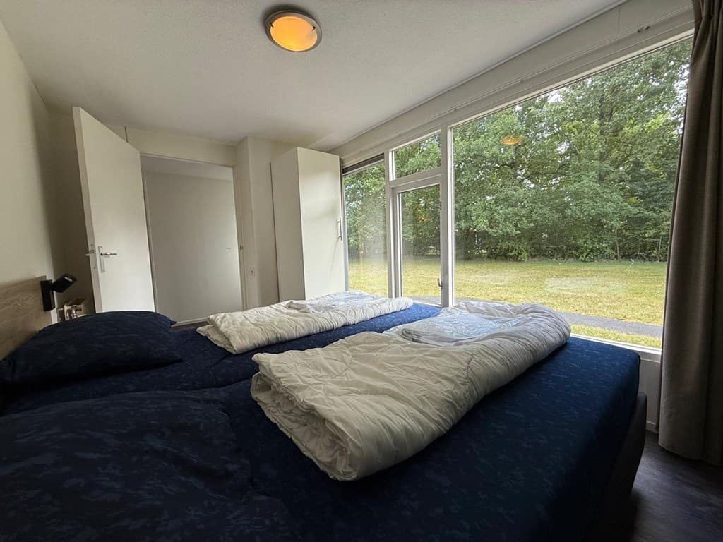 Slaapkamer met twee bedden en grote ramen met uitzicht op een groene tuin en bomen.