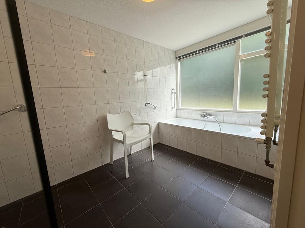 Lichte en ruime badkamer met ligbad, aparte douche en een witte stoel bij een groot raam
