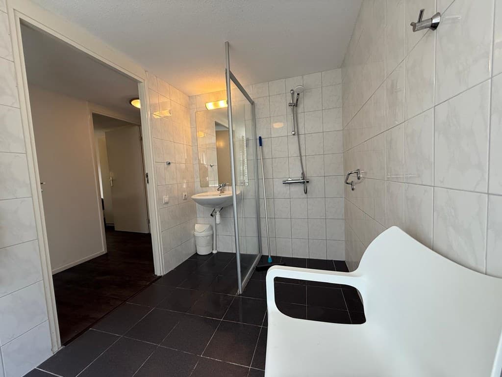 Ruime badkamer met wastafel, douchecabine en een witte bank in de hoek, betegelde wanden en vloeren.