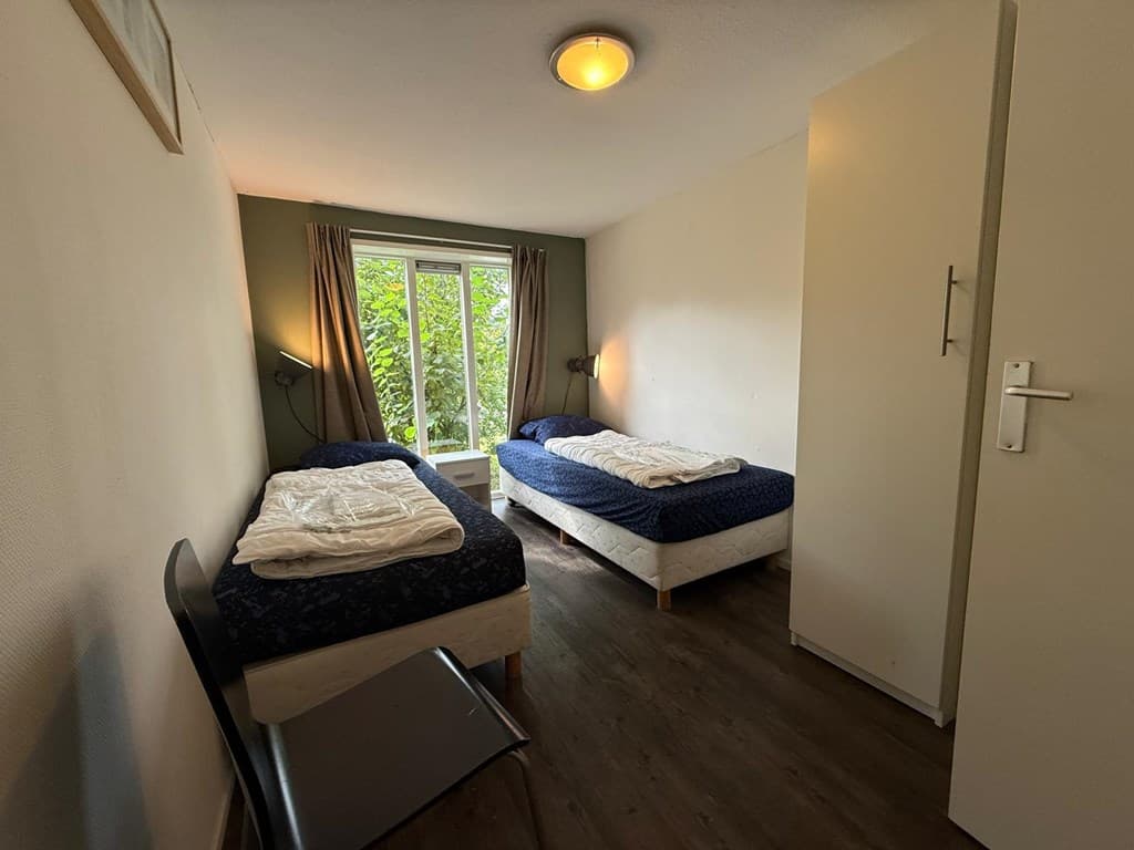 Slaapkamer met twee éénpersoonsbedden, een stoel en raam met uitzicht op groen.