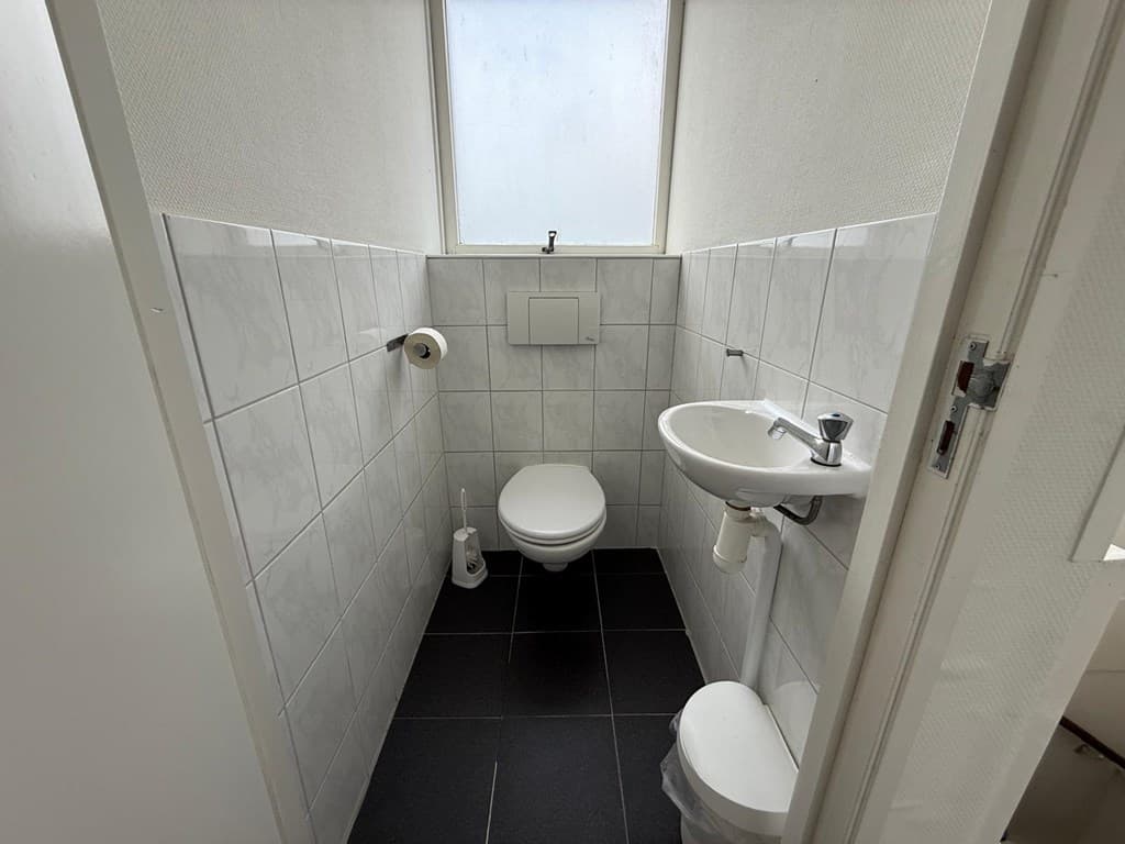 Verlichting toiletruimte met witte tegels, toilet en wastafel bij een raam met natuurlijke lichtinval