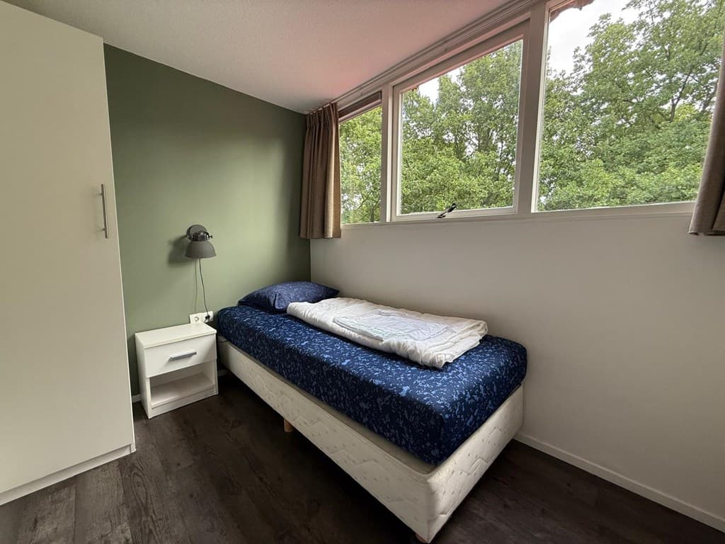 Een slaapkamer met een eenpersoonsbed, nachtkastje en grote ramen met uitzicht op groene bomen.