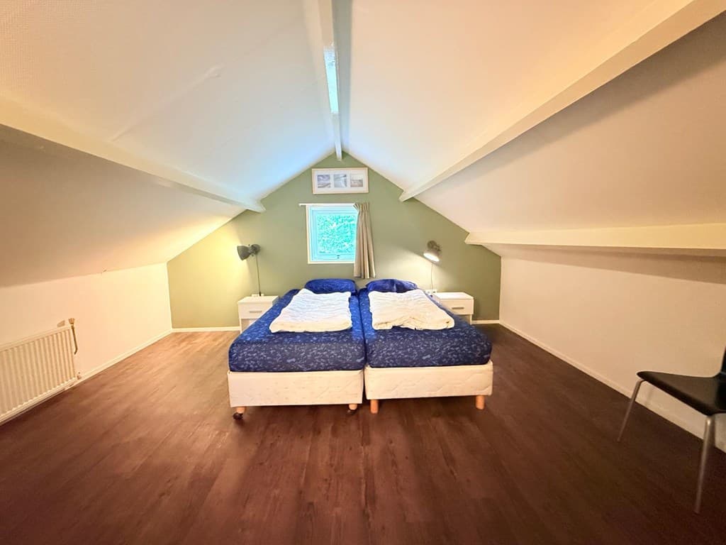 Ruimtelijke slaapkamer met twee bedden, houten vloer en schuin plafond, voorzien van een raam en nachtkastjes.