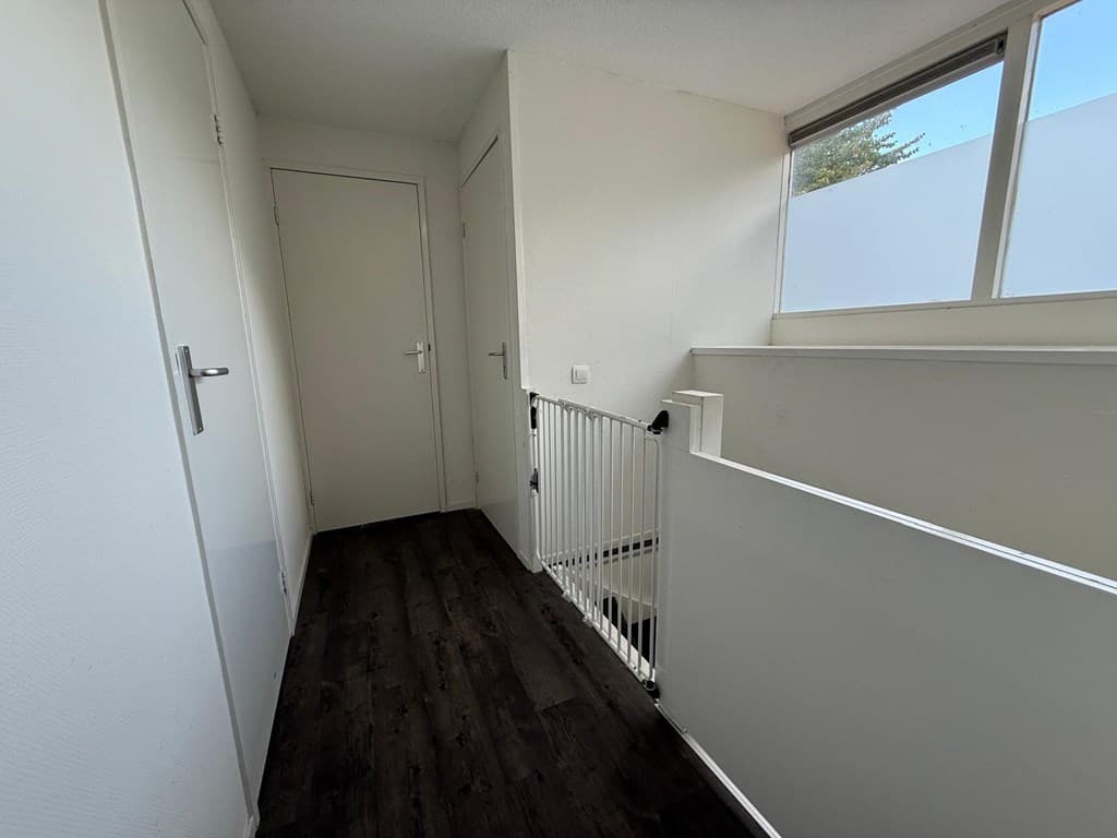 Lichte hal met laminaatvloer, witte wanden en een wand met een reling naar beneden. Deuren aan beide zijden zichtbaar.