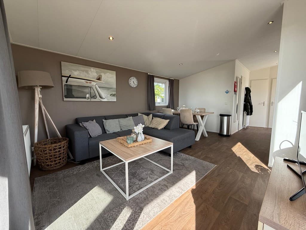 Woonruimte met een grijze bank, houten tafel, vloerkleed en grote ramen die natuurlijk licht binnenlaten