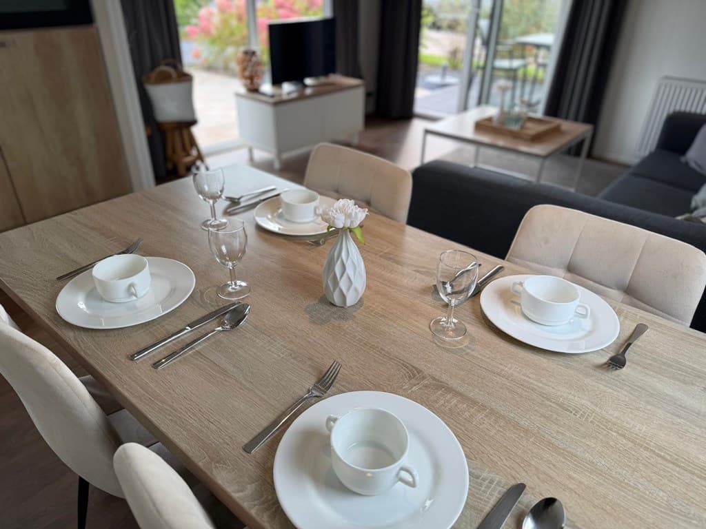 Eettafel met witte borden, bestek en glazen, met op de achtergrond een moderne woonkamer en uitzicht op de tuin.