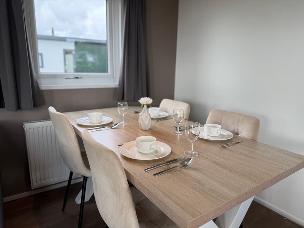 Eettafel met vier stoelen, gedekt met borden en glazen, en een vaasje met een bloem bij een raam.