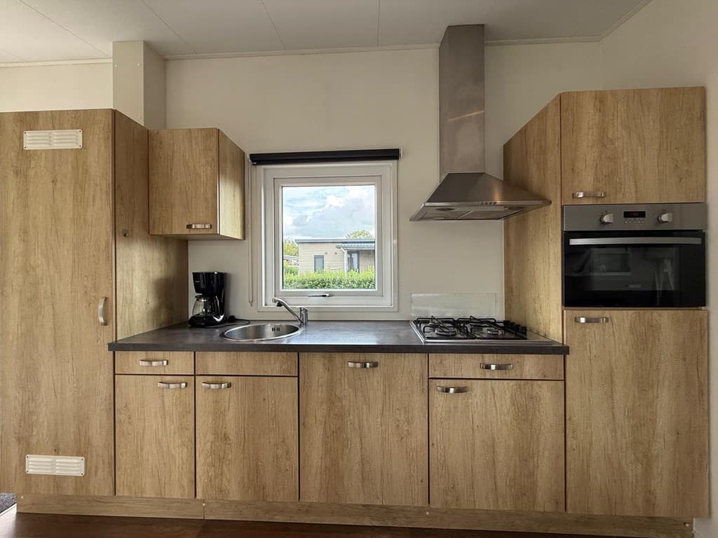 Keuken met houten kasten, kookplaat, oven, spoelbak en raam met uitzicht op de buitenruimte.