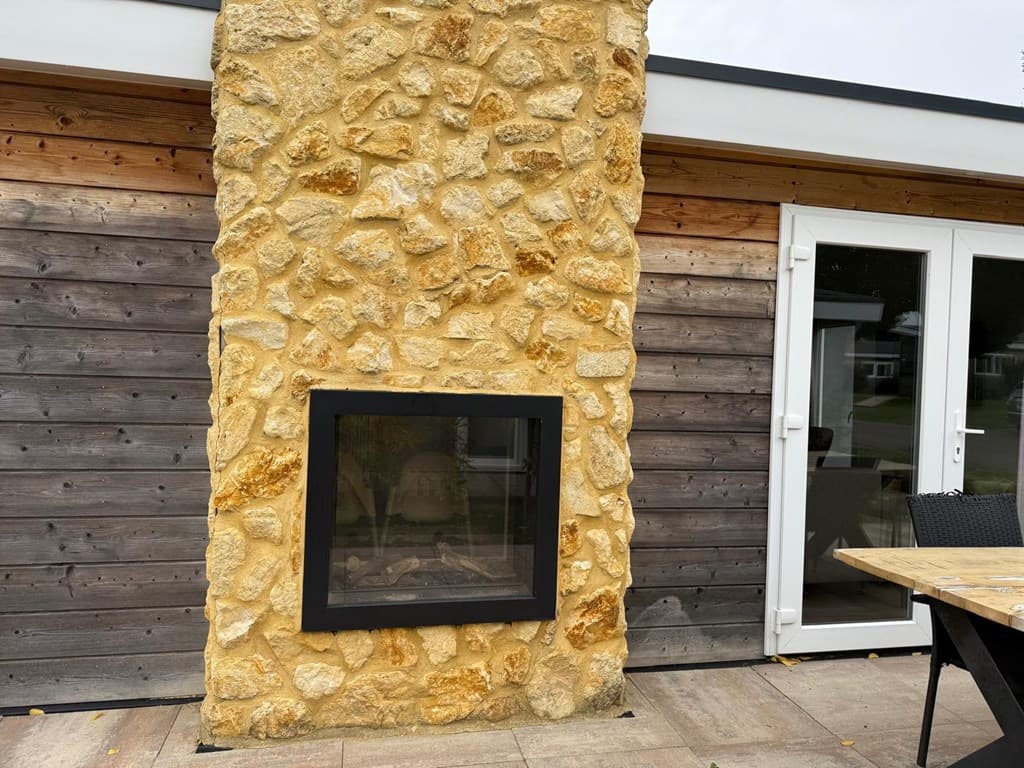 Buitenzijde van een woning met een stenen gevel en een moderne inbouwhaard naast een houten wand.