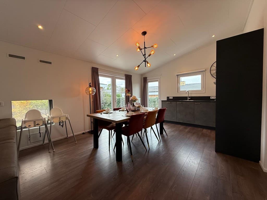 Eetkamer met een houten tafel en vier stoelen, grote ramen en moderne keuken met donkere kasten
