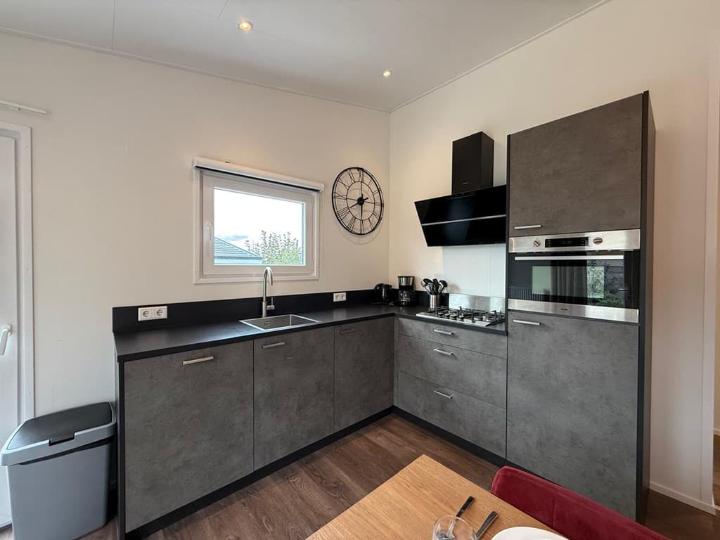 Moderne keuken met grijze kasten, spoelbak, kookplaat en oven, naast een raam met uitzicht naar buiten.