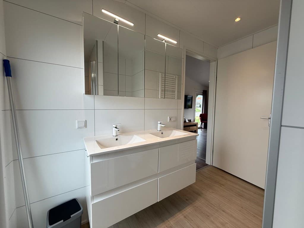 Badkamer met dubbele wastafel, grote spiegel en lichte wandafwerking, toegang tot andere ruimtes zichtbaar.
