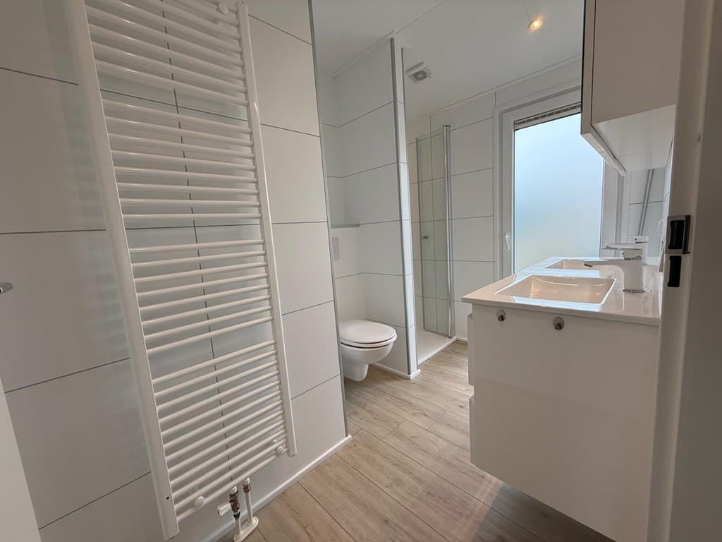 Moderne badkamer met witte tegels, wastafels, toilet en een inloopdouche met glazen wand