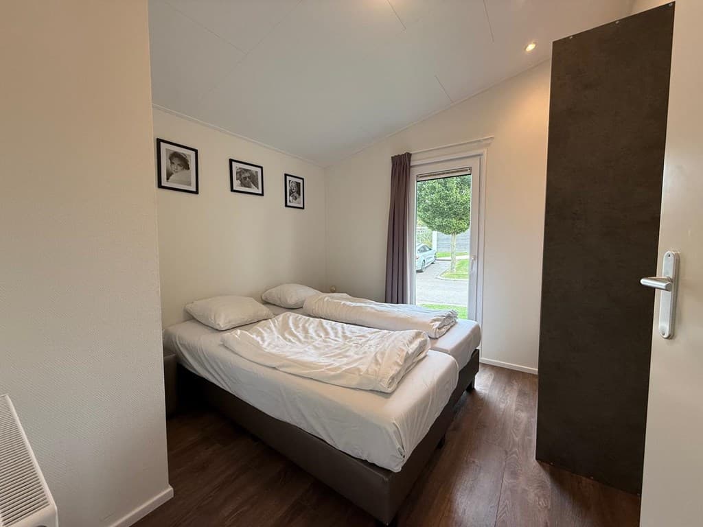 Slaapkamer met twee eenpersoonsbedden, raampartij met gordijnen en houten vloer, enkele fotolijsten aan de muur.