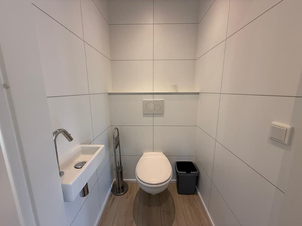 Moderne toiletruimte met witte tegels, een wandtoilet, een fonteintje en een prullenbak.
