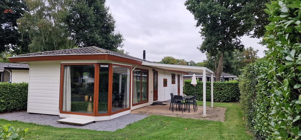 Moderne vakantiewoning met grote ramen, een terras en tuinmeubelen, omringd door groene hagen en bomen