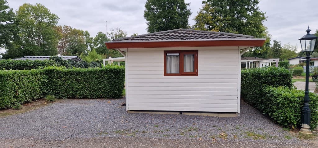 Wit vakantiehuisje met een houten dak, omringd door heggen en een grindpad voor de ingang