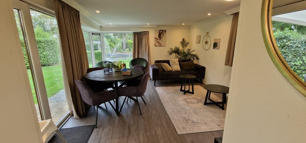 Moderne woonkamer met grote ramen, ronde eettafel en comfortabele zithoek met bank en planten.