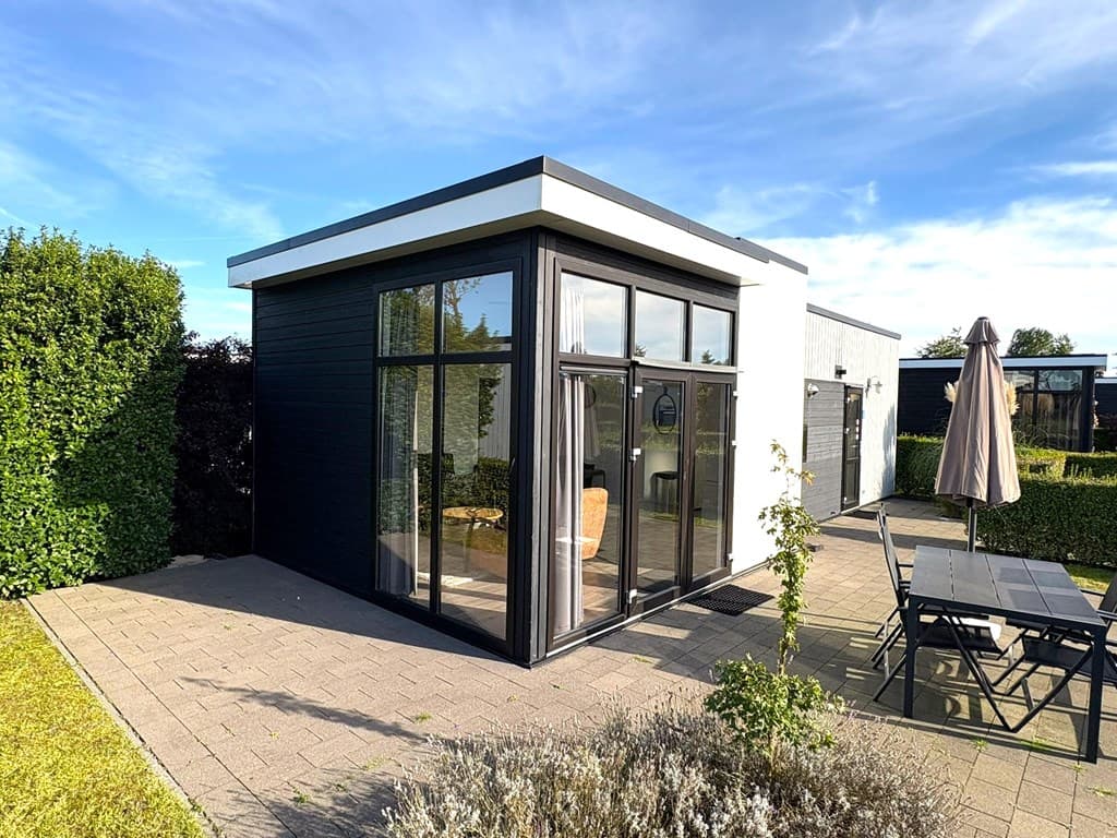Moderne vakantiewoning met grote ramen en een terras, omgeven door groen en een bestrate oprit.