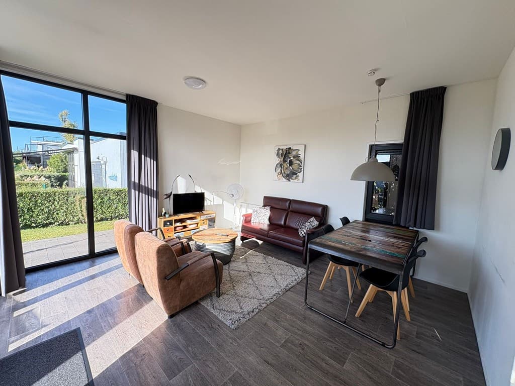 Moderne woonkamer met zithoek, eettafel en grote ramen met zicht op buitenruimte en groen.