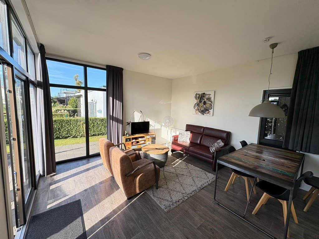 Interieur van een vakantiehuis met zithoek, eettafel en grote ramen met uitzicht op groen.