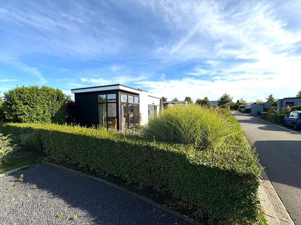Moderne vakantiewoning met grote ramen, omgeven door groen en grindpad, gelegen in een rustige omgeving.